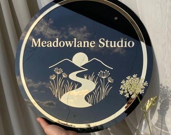 Puede incluir: Letrero redondo negro con borde dorado y texto "Meadowlane Studio". El diseño incluye un paisaje con montañas, un sol y un río. El letrero está sostenido por una persona.