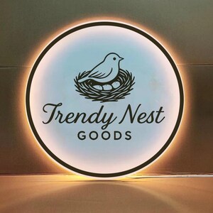 Może przedstawiać: Okrągły, podświetlany znak z czarną obw&oacute;dką. Znak przedstawia ptaka w gnieździe z jajkami oraz napis "Trendy Nest GOODS" w ozdobnej czcionce. Tło to gradient jasnoniebieskiego i białego.