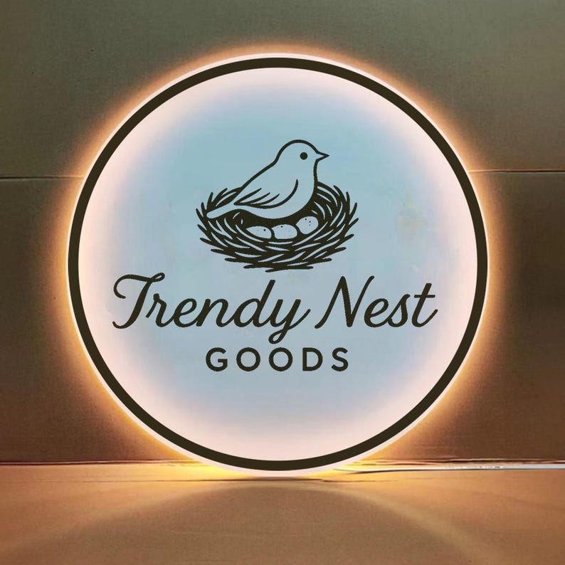 Może przedstawiać: Okrągły, podświetlany znak z czarną obw&oacute;dką. Znak przedstawia ptaka w gnieździe z jajkami oraz napis "Trendy Nest GOODS" w ozdobnej czcionce. Tło to gradient jasnoniebieskiego i białego.