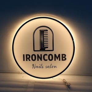 May include: A round, illuminated sign with a black border. The sign features a white background with a black comb logo inside a rounded rectangle. The text "IRONCOMB Nails salon" is displayed in black, with a warm glow emanating from the sign.