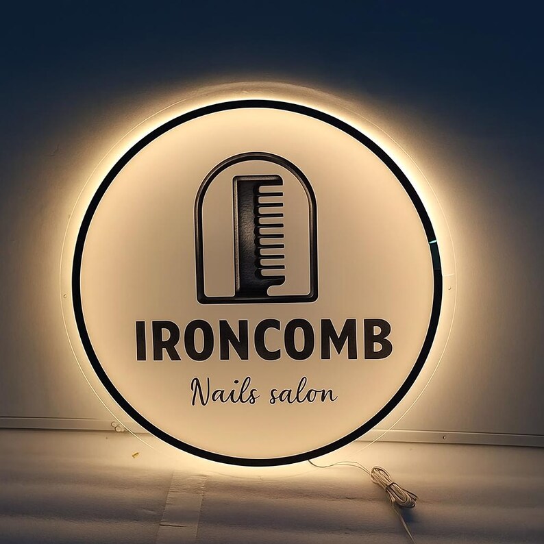 May include: A round, illuminated sign with a black border. The sign features a white background with a black comb logo inside a rounded rectangle. The text "IRONCOMB Nails salon" is displayed in black, with a warm glow emanating from the sign.