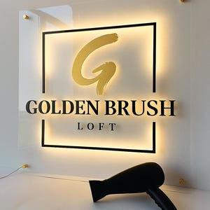 Może przedstawiać: Podświetlany szyld dla Golden Brush Loft, z złotą literą 'G' i tekstem. Znak jest zamontowany na przezroczystym panelu ze złotym osprzętem. Czarna suszarka do włos&oacute;w spoczywa poniżej.