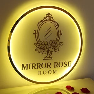 Może przedstawiać: Okrągły, podświetlany znak ze złotą obw&oacute;dką i napisem "MIRROR ROSE ROOM". Znak zawiera dekoracyjne lustro i wz&oacute;r r&oacute;ży. Czerwone płatki r&oacute;ż są rozrzucone poniżej.
