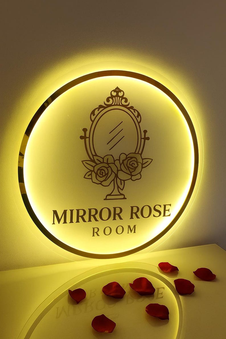 Może przedstawiać: Okrągły, podświetlany znak ze złotą obw&oacute;dką i napisem "MIRROR ROSE ROOM". Znak zawiera dekoracyjne lustro i wz&oacute;r r&oacute;ży. Czerwone płatki r&oacute;ż są rozrzucone poniżej.
