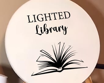 Puede incluir: Un letrero redondo blanco con texto negro que dice "LIGHTED Library" y una silueta negra de un libro abierto.