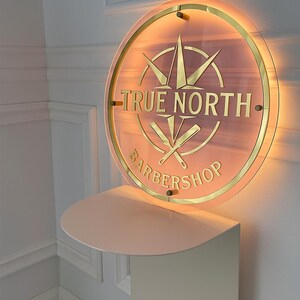 May include: A round, illuminated sign for a barbershop. The sign features the words "TRUE NORTH BARBERSHOP" in gold lettering, along with a compass design and crossed barber tools. The sign is mounted on a light pink shelf.