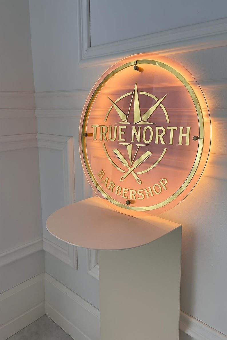 May include: A round, illuminated sign for a barbershop. The sign features the words "TRUE NORTH BARBERSHOP" in gold lettering, along with a compass design and crossed barber tools. The sign is mounted on a light pink shelf.