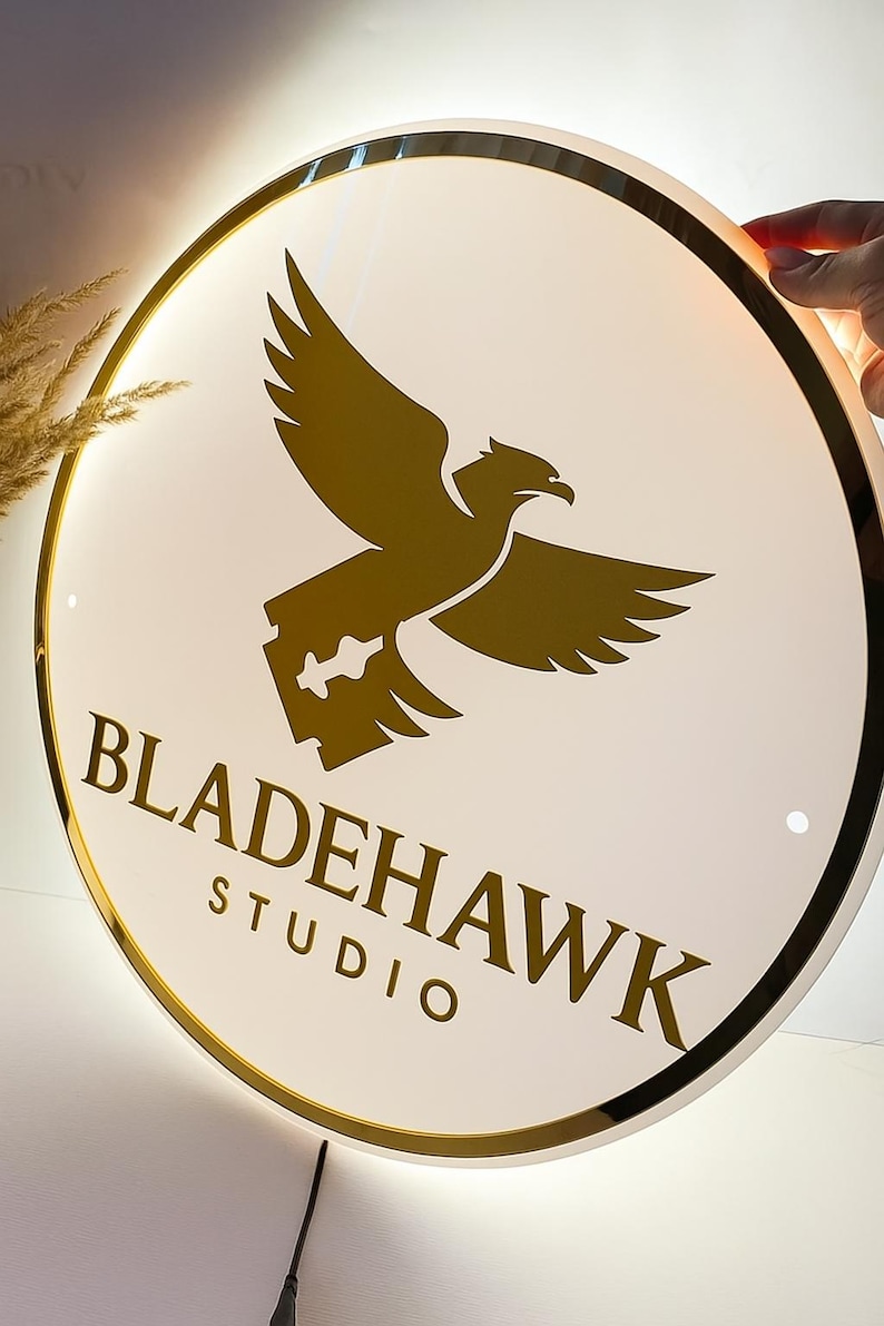 May include: Round, illuminated sign with a gold border and a white face. The sign features a gold eagle graphic and the text "BLADEHAWK STUDIO" in a serif font. The sign is lit from behind.