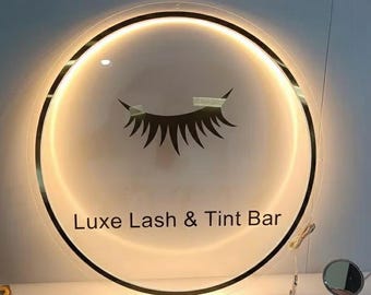 Puede incluir: Un letrero redondo e iluminado con un borde negro y el texto "Luxe Lash & Tint Bar". El letrero presenta un diseño estilizado de pestañas. El letrero está iluminado con un brillo cálido y acogedor.