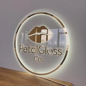 May include: A round, illuminated sign with a gold-colored border and the words "PetalGloss Co." in a gold font. The sign features a gold lip design in the center. The sign is mounted on a white wall.