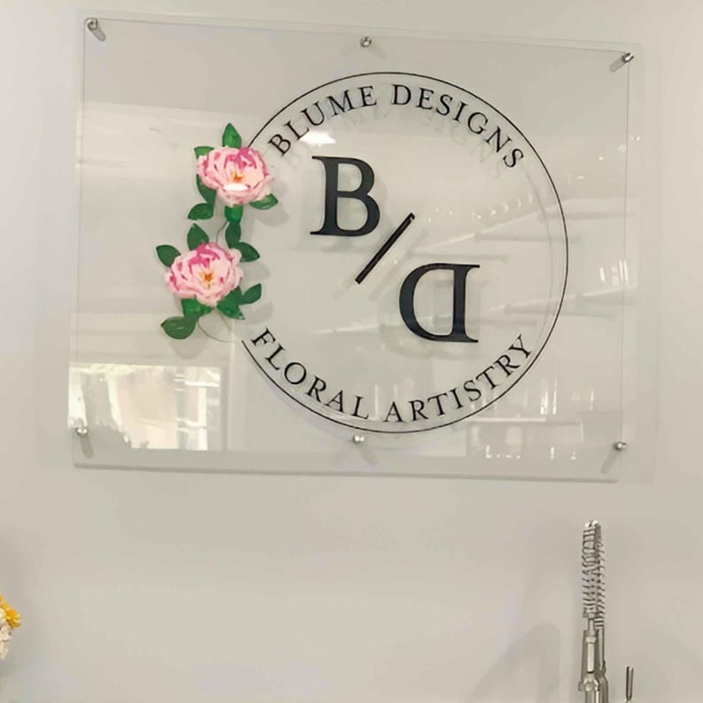 May include: A clear acrylic sign with a black circle containing the text "Blume Designs B/D Floral Artistry". Two pink paper flowers are attached to the left side of the circle.