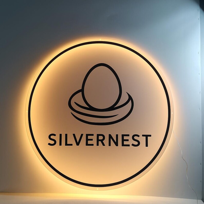 May include: Circular sign with a glowing edge. The sign features a black outline of an egg in a nest above the word "SILVERNEST" in a bold, black font. The background is a soft, off-white color.
