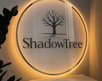 Puede incluir: Un letrero redondo e iluminado con el texto "ShadowTree" en negro, con una fuente elegante. El letrero presenta un diseño de árbol silueteado y una luz cálida y brillante alrededor del borde. El letrero está montado en una pared.