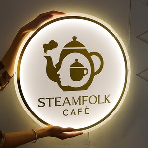 May include: A round, illuminated sign with a gold-colored border and a white center. The sign features a gold-colored teapot design with a face silhouette and a teacup. The text "STEAMFOLK CAF&Eacute;" is displayed in gold.