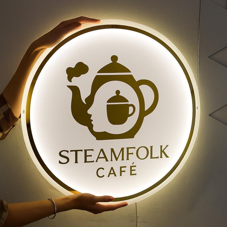 May include: A round, illuminated sign with a gold-colored border and a white center. The sign features a gold-colored teapot design with a face silhouette and a teacup. The text "STEAMFOLK CAF&Eacute;" is displayed in gold.