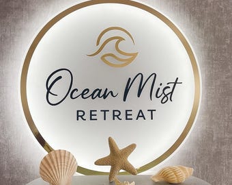 May include: A round, illuminated sign with the words "Ocean Mist Retreat" in elegant script. The sign features a gold wave design and a gold border. Seashells and starfish are arranged on a gray surface in front of the sign.