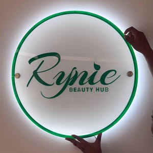 May include: A round, illuminated sign with a green border and the text "Rynie BEAUTY HUB" in green script. The sign is white with a bright, glowing light around the edges. Two small, round fasteners are visible.