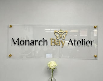 Puede incluir: Letrero rectangular transparente con las palabras "Monarch Bay Atelier" en letras negras y doradas. Un logotipo de mariposa dorada está encima de la palabra "Atelier". El letrero está montado en una pared con tornillos dorados. Una rosa blanca en un jarrón de cristal está en primer plano.