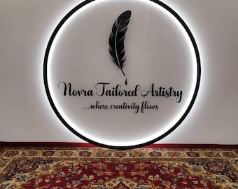 Puede incluir: Un letrero circular con un borde negro y un interior blanco iluminado. El letrero presenta un diseño de pluma negra y el texto "Novra Tailored Artistry ...where creativity flows". Una alfombra estampada está en primer plano.