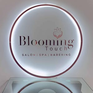 May include: A circular sign with the words "Blooming Touch" in a stylized font. The sign has a glowing white interior and a rose gold border. Below the main text are the words "Salon | Spa | Babering".