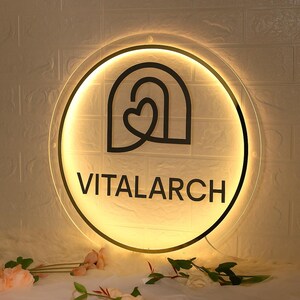 May include: A round, illuminated sign with the word "VITALARCH" in black, centered below a black graphic of a heart within an arch. The sign has a warm, yellow glow and is mounted on a white brick wall. Decorative flowers are in the foreground.