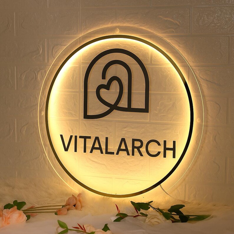 May include: A round, illuminated sign with the word "VITALARCH" in black, centered below a black graphic of a heart within an arch. The sign has a warm, yellow glow and is mounted on a white brick wall. Decorative flowers are in the foreground.