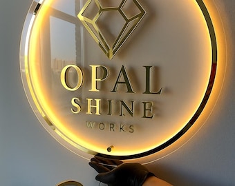Puede incluir: Un letrero redondo e iluminado con un logotipo de diamante dorado y el texto "OPAL SHINE WORKS". El letrero tiene un borde dorado y una luz cálida y brillante. Un pequeño espejo compacto dorado está en primer plano.