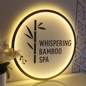 Może przedstawiać: Okrągły, podświetlany znak z ramą w kolorze złota. Znak przedstawia czarny wz&oacute;r bambusa i słowa "WHISPERING BAMBOO SPA" w nowoczesnej czcionce. Tło jest miękkie, świecące na biało.