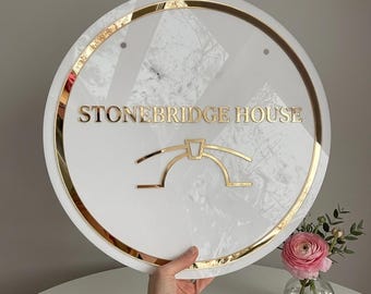 Puede incluir: Un letrero circular con fondo de patrón de mármol blanco y borde dorado. El texto "STONEBRIDGE HOUSE" está centrado en letras doradas sobre un icono de puente dorado. El letrero tiene dos pequeños orificios en la parte superior para montaje.