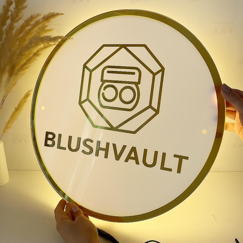 May include: A round, illuminated sign with a gold border and the text "BLUSHVAULT" in a bold, sans-serif font. The sign features a gold geometric design with a makeup compact icon in the center.