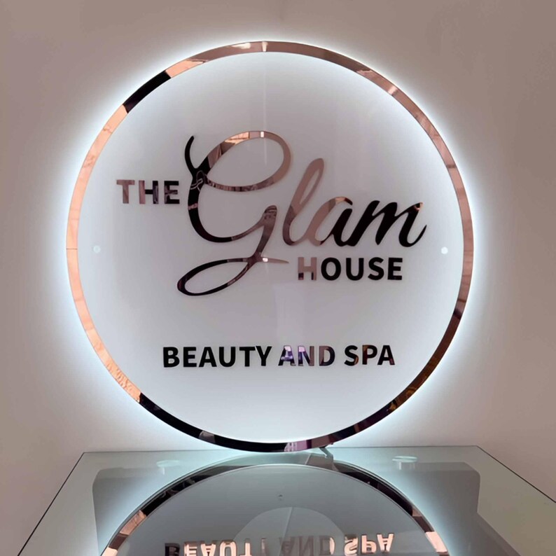 May include: A round, illuminated sign with a rose gold trim and the words "THE Glam HOUSE BEAUTY AND SPA" in a stylish font. The sign is mounted on a clear surface, reflecting the text below.