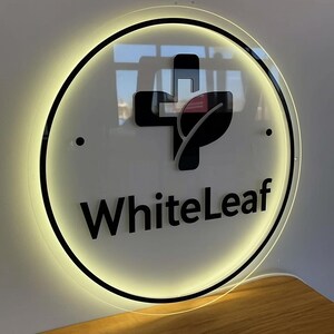 Peut inclure: Panneau circulaire avec une bordure noire et les mots "WhiteLeaf" en gras. Le panneau pr&eacute;sente un motif de croix et de feuille noir. Le panneau est &eacute;clair&eacute; par une lumi&egrave;re jaune chaude.