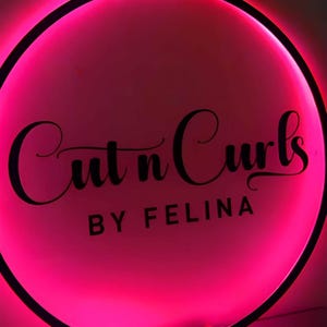 May include: A round, illuminated sign with a vibrant pink glow. The words "Cut n Curls" are written in elegant cursive script, with "BY FELINA" below in a sans-serif font. The sign has a black border.