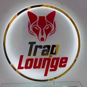 May include: A round, illuminated sign with a red fox head logo above the words "Trap Lounge" in red and black lettering. The sign has a gold-colored border and a white background, creating a striking contrast.