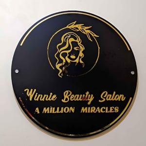 May include: A black circular sign with gold lettering and a gold silhouette of a woman with long hair and an olive branch. The sign reads "Winnie Beauty Salon, A Million Miracles".