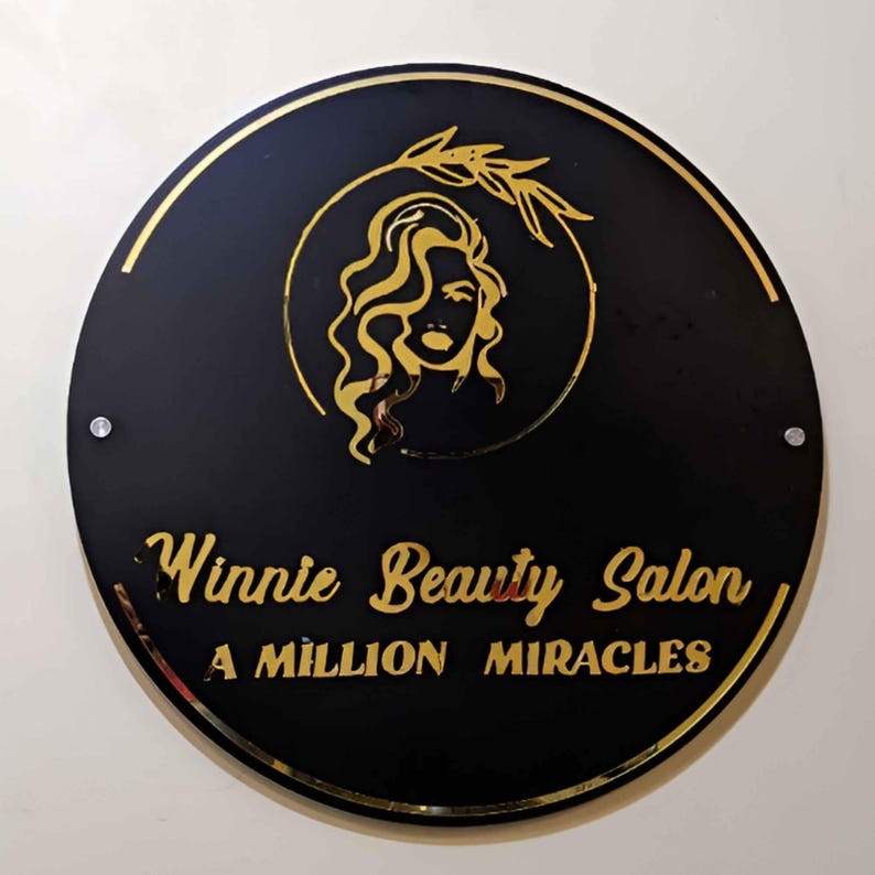 May include: A black circular sign with gold lettering and a gold silhouette of a woman with long hair and an olive branch. The sign reads "Winnie Beauty Salon, A Million Miracles".