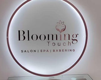 Puede incluir: Un letrero redondo e iluminado con las palabras "Blooming Touch" en una fuente elegante. El letrero también incluye el texto "SALON | SPA | BABERING" y un logotipo floral. Tiene un borde de oro rosa y está montado sobre una superficie de vidrio.