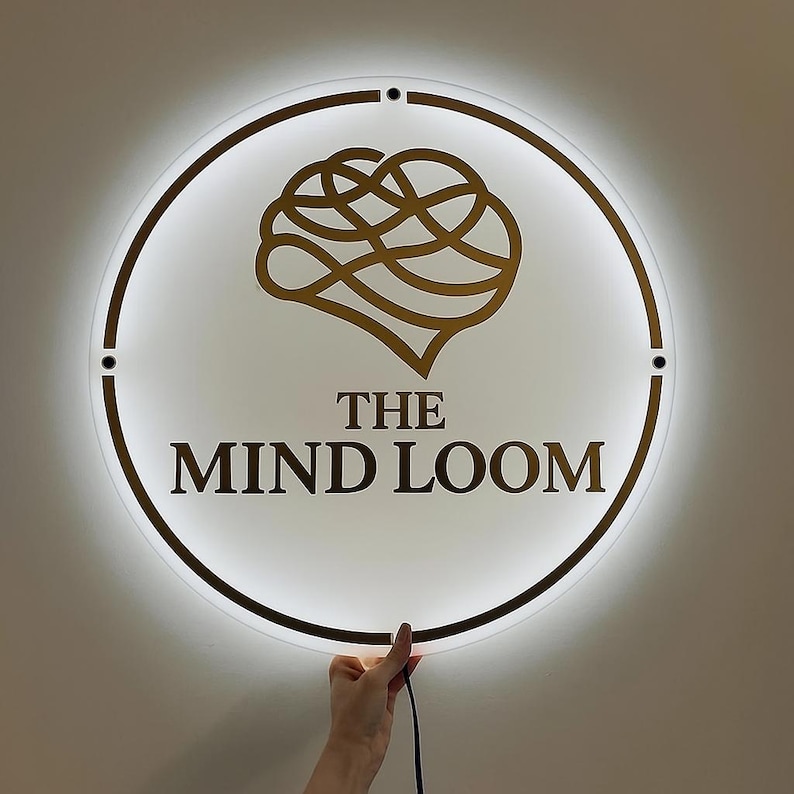May include: A round, illuminated sign with a gold brain graphic and the text "THE MIND LOOM" in a bold, sans-serif font. The sign has a white background and a gold border, with a hand holding the power cord.