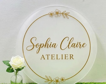 Puede incluir: Un letrero circular de acrílico esmerilado con el texto dorado Sophia Claire ATELIER en el centro. El letrero está decorado con un borde circular dorado fino y arte lineal floral dorado en la parte superior e inferior. Una rosa blanca está en un florero.