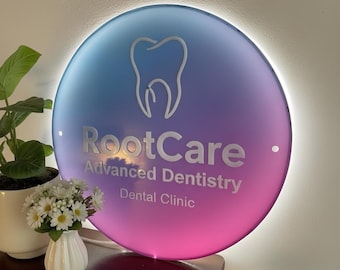 Puede incluir: Un letrero redondo con un degradado de azul, morado y rosa, con el texto "RootCare Advanced Dentistry Dental Clinic" y un gráfico de un diente. El letrero está iluminado y montado en una pared.
