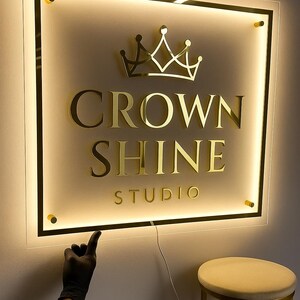 Może przedstawiać: Podświetlany szyld dla studia, z napisem "CROWN SHINE STUDIO" złotymi literami i złotym emblematem korony. Znak jest zamknięty w czarnej ramie z ciepłym, świecącym podświetleniem. Ręka w czarnej rękawicy wskazuje na znak.