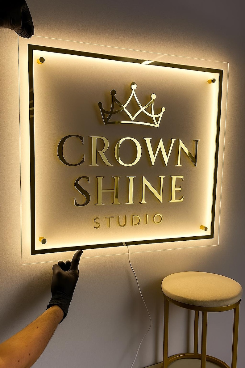 Może przedstawiać: Podświetlany szyld dla studia, z napisem "CROWN SHINE STUDIO" złotymi literami i złotym emblematem korony. Znak jest zamknięty w czarnej ramie z ciepłym, świecącym podświetleniem. Ręka w czarnej rękawicy wskazuje na znak.