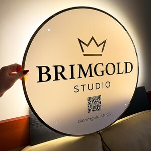 May include: A round, illuminated sign with a gold-colored trim. The sign features the text "BRIMGOLD STUDIO" in bold black letters, a crown graphic, and a QR code. The sign also includes the Instagram handle "@brimgold_studio".