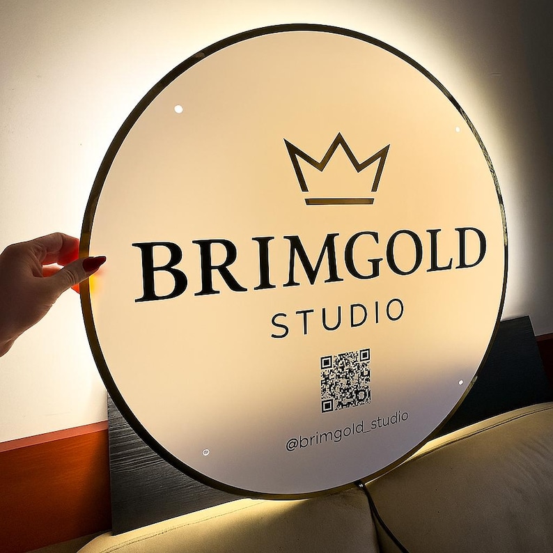 May include: A round, illuminated sign with a gold-colored trim. The sign features the text "BRIMGOLD STUDIO" in bold black letters, a crown graphic, and a QR code. The sign also includes the Instagram handle "@brimgold_studio".