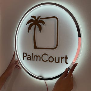 May include: A round, illuminated sign with a brown palm tree graphic and the text "PalmCourt". The sign has a silver border and a white background. The sign is being held by two hands.