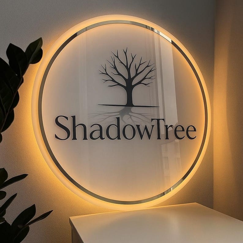 May include: A round, illuminated sign with the text "ShadowTree" in a black, elegant font. The sign features a silhouetted tree design and a warm, glowing light around the edge. The sign is mounted on a wall.