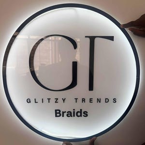May include: A round, illuminated sign with a black border and the text "GT" in large, black letters. Below, the words "GLITZY TRENDS" and "Braids" are displayed in a smaller, black font against a white background.