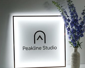 Puede incluir: Un letrero cuadrado e iluminado con el texto "Peakline Studio" en una fuente moderna. El letrero presenta un logotipo negro encima del texto y está rodeado por un marco delgado y oscuro. Un jarrón con flores azules está a la derecha.