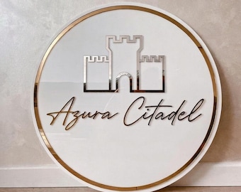 Puede incluir: Un letrero redondo blanco con un borde dorado y las palabras "Azura Citadel" en una fuente cursiva. El letrero presenta un emblema de castillo plateado en el centro.