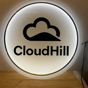 May include: Round, illuminated sign with a white background and a black cloud logo. The text "CloudHill" is displayed in black, centered below the logo. The sign has a gold-colored trim and is lit from within.